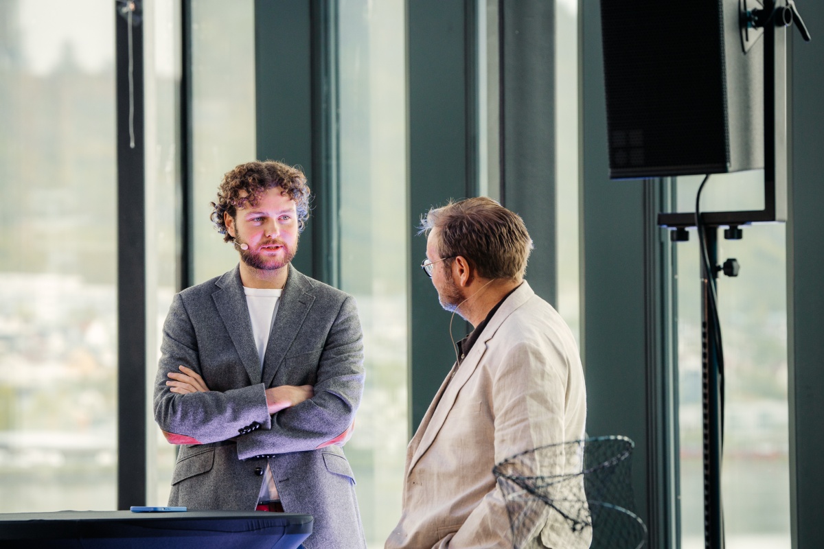 To menn står på en scene og samtaler med hverandre under en konferanse. Begge har mikrofoner påmontert, og bak dem skimtes store vinduer med utsikt over et bylandskap. 