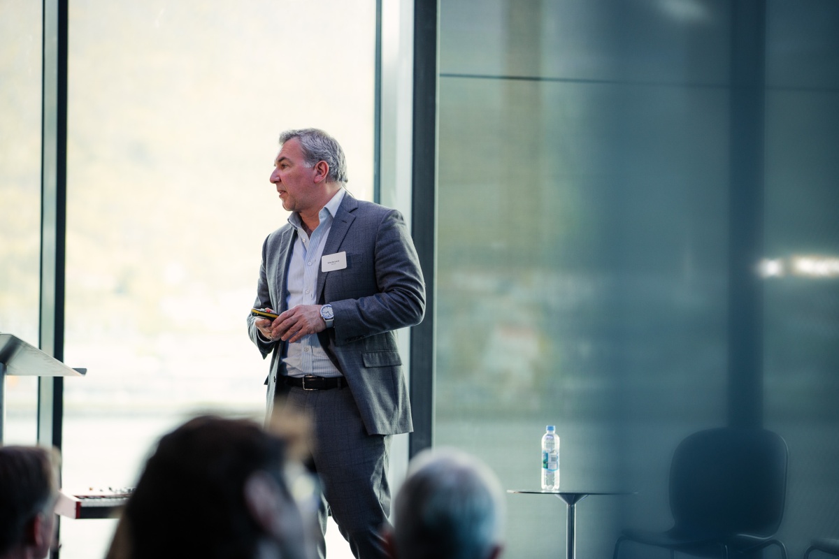 En mann i grå dress står på scenen og holder et innlegg. Han ser mot venstre og holder en presentasjonsklikker i hånden. Publikum skimtes i forgrunnen, og store vinduer bak gir utsyn mot fjell og hav.