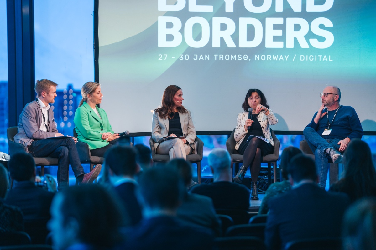 Et panel med fem personer sitter på en scene foran et publikum under Arctic Frontiers 2025, med et stort bakskjermbanner som viser teksten “Beyond Borders” og utsikt over fjell og hav gjennom de store vinduene i bakgrunnen.