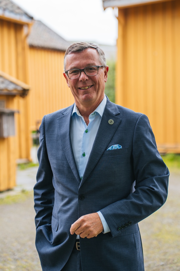 Smilende mann i dress og briller står utendørs mellom gule trebygninger.