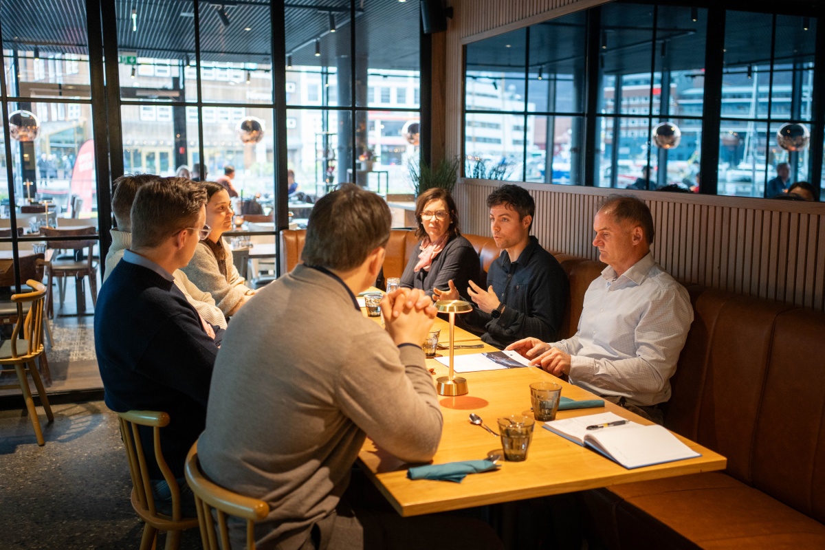 Seks personer sitter rundt et bord på en kafé og deltar i en samtale. På bordet ligger notatbøker, dokumenter og vannglass.