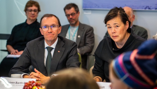 To møtedeltakere sitter ved et bord med navneskilt og frukt, mens flere andre følger med i bakgrunnen under et formelt møte.