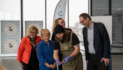 En kokk viser frem mat til en gruppe smilende personer under et arrangement, med bannere i bakgrunnen.