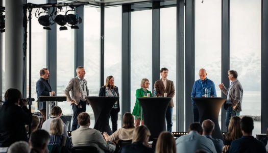 Syv paneldeltakere står samlet bak høye bord på en scene under en konferanse, med et publikum foran seg. Store vinduer i bakgrunnen viser snødekte fjell og hav. En deltaker til høyre snakker mens de andre lytter.