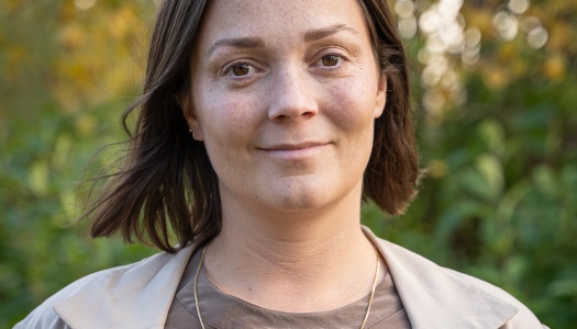 Portrett av en kvinne utendørs, med kort brunt hår, beige jakke og brun topp, foran grønn og gyllen høstlig bakgrunn.