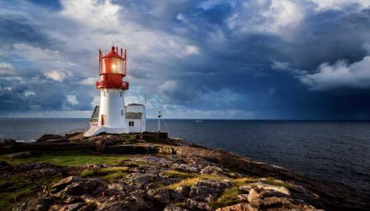 Fyrtårn med hvit base og rød topp står på en klippe ved havet, med dramatisk himmel og skyer i bakgrunnen.