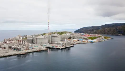 Oversiktsbilde av et stort industrikompleks ved kysten, med røyksøyle fra en pipe og fjell i bakgrunnen.