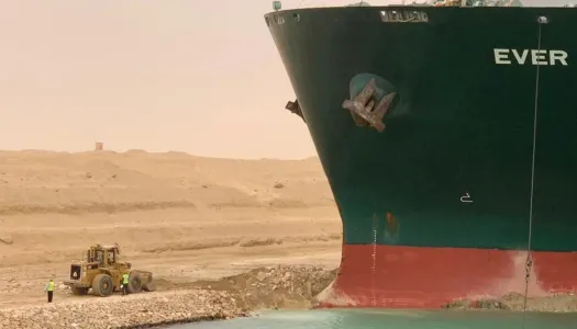 Et stort containerskip sitter fast på grunn i en kanal, mens en gravemaskin og arbeidere står ved siden av for å prøve å frigjøre det.