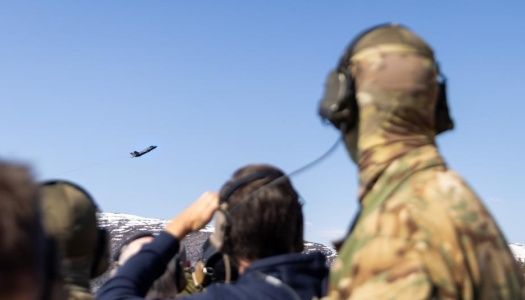 oldater med hørselvern og kamuflasjeuniform ser på et jagerfly som flyr over et fjellområde under en militær øvelse.