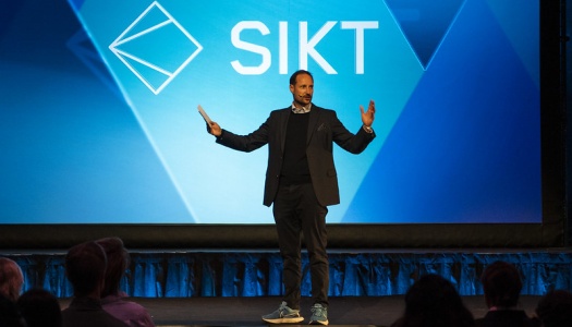 En mann står på scenen foran et stort lerret med teksten «SIKT». Han snakker til publikum med åpne armer.