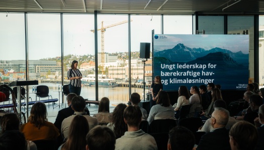En kvinne står og holder innlegg foran et publikum i et lyst lokale med store vinduer mot havna i Tromsø. På en stor skjerm ved siden av henne står teksten «Ungt lederskap for bærekraftige hav- og klimaløsninger», med logoen til Senter for hav og Arktis. Publikum følger med på presentasjonen, og bakgrunnen viser bygg, båter og en kran ved havna.