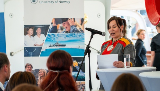 En kvinne i en rød, grå og oransje kofte står foran en mikrofon og taler til en forsamling. 