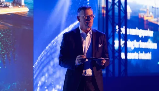 Mann i dress står på en scene med blått lys og store skjermer i bakgrunnen, og holder talekort mens han snakker til publikum under en konferanse.