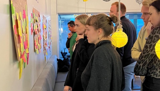 En gruppe deltakere står samlet foran en vegg med store ark dekket av fargerike post-it-lapper, og diskuterer ideer i en workshop-setting. Stemningen er konsentrert og engasjert