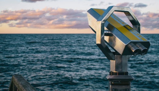 En kikkert montert på en brygge vender utover havet ved solnedgang, med rosa skyer og blått hav i horisonten.