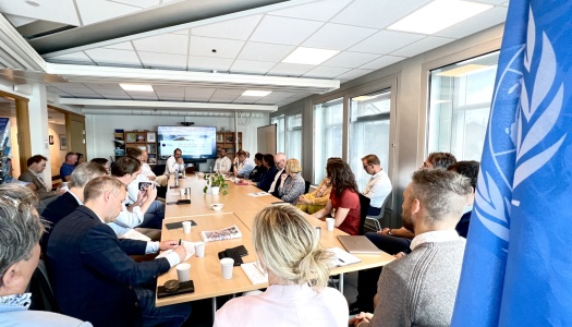 Møte i konferanserom med mange deltakere samlet rundt et langt bord, presentasjon vises på skjerm i enden av rommet, med en FN-flagg synlig i forgrunnen.