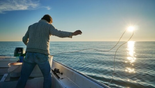 En person står i en liten båt og kaster en line ut i havet mens solen står lavt over horisonten. Sollyset glitrer i det rolige vannet og skaper en varm stemning i kveldssolen.