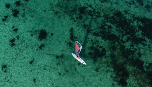 Luftfoto av en person som seiler på et brett med fargerikt seil over grunt, turkist vann. Havbunnen med mørke flekker av tang og stein skimtes tydelig gjennom det klare vannet.