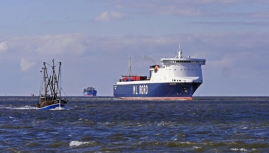 Et stort lasteskip med teksten "ML RORO" på skroget seiler på havet, mens en mindre fiskebåt passerer i forgrunnen. I horisonten skimtes flere fartøy under en delvis skyet himmel.
