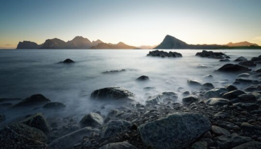Kystlandskap i solnedgang med steiner i forgrunnen og fjell i horisonten. Lang eksponering gir havet et sløret, tåkete uttrykk som kontrasterer mot de mørke fjellformasjonene i bakgrunnen.