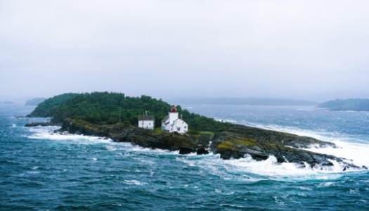 En liten øy med et hvitt fyrtårn og bolighus omgitt av grønn vegetasjon, ligger ute i et opprørt hav. Bølgene slår mot klippene, og tåke og gråvær gir landskapet et dramatisk kystpreg.