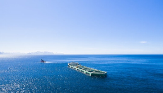 En oppdrettsmerd slepes utover havet av en båt under klar blå himmel. Det dype blå vannet strekker seg mot horisonten, der fjellene skimtes i det fjerne.