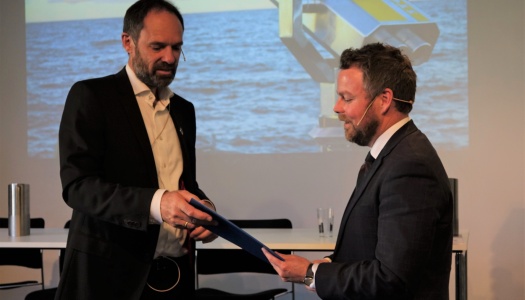 To menn i dress står på en scene under en presentasjon, der den ene overrekker et dokument til den andre. I bakgrunnen vises et bilde av havet og en kikkert på storskjerm.