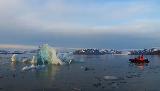 Et isfjell flyter i rolig arktisk farvann med snødekte fjell i bakgrunnen. En liten gummibåt med personer i oransje redningsdrakter nærmer seg isfjellet under kveldssolens varme lys, som speiler seg i vannflaten.