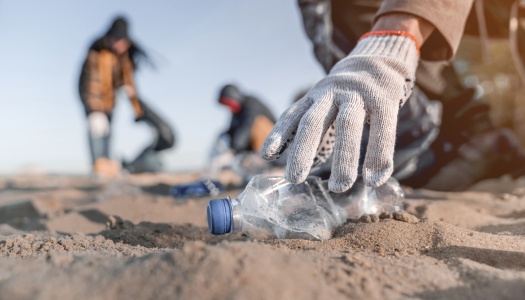 Hånd med hanske plukker opp en plastflaske fra sanden under strandrydding, med flere personer som rydder i bakgrunnen.