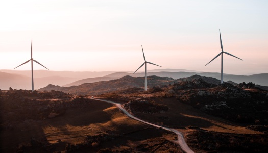 re vindmøller står på en åsrygg i et landskap med fjell og daler, badet i varmt kveldslys.