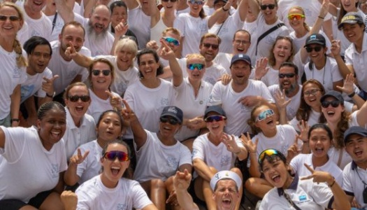Stort gruppebilde av mennesker i hvite t-skjorter på dekket av et seilskip, smilende og hevende armene i en begeistret pose.