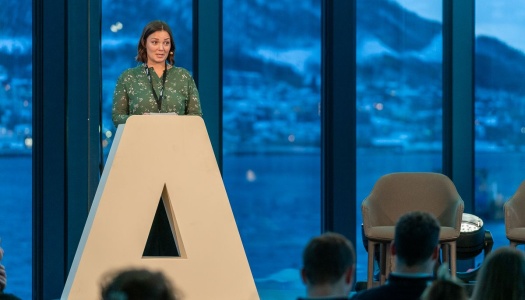 En person står på en scene bak en stor talerstol formet som en “A”, og holder et innlegg foran et publikum, med vinterlandskap og fjell synlig gjennom store vinduer i bakgrunnen.