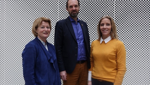 Tre personer står foran en hvit vegg med sirkulære hull. De smiler mot kameraet; to kvinner på hver side og en mann i midten.