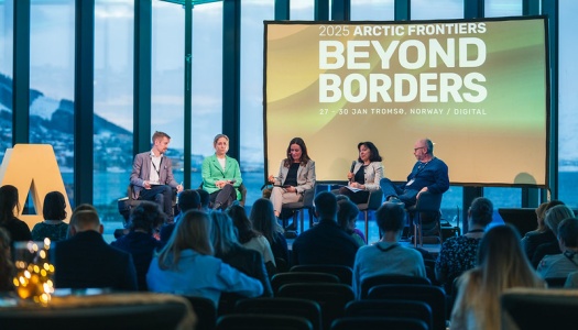 Et panel med fem personer sitter på en scene foran et publikum under Arctic Frontiers 2025, med et stort bakskjermbanner som viser teksten “Beyond Borders” og utsikt over fjell og hav gjennom de store vinduene i bakgrunnen.