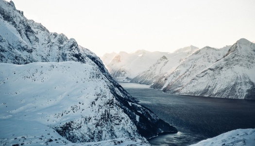 Panoramisk vinterlandskap med en smal fjord som snor seg mellom høye, snødekte fjell under en blek himmel. Solen kaster et mykt lys på fjelltoppene, og mørkeblått vann kontrasterer mot de hvite fjellsidene.