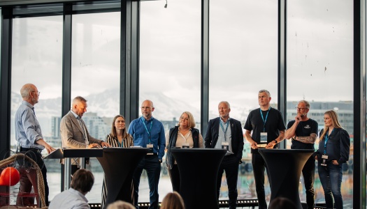 Et panel med ni personer fra ulike deler av sjømatnæringen og forvaltningen står samlet foran store vinduer med utsikt mot fjell og by. De deltar i en panelsamtale foran publikum under en konferanse.