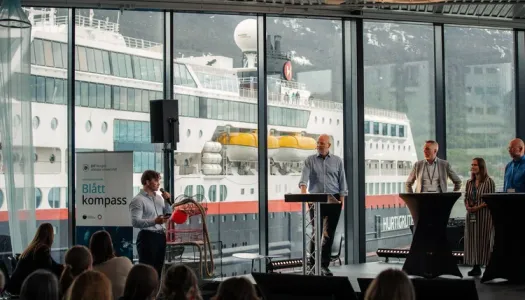 En mann står med mikrofon og snakker til fire mennesker på scenen foran et publikum under arrangementet Blått Kompass.Et Hurtigruten-skip er synlig gjennom de store panoramavinduene i bakgrunnen.