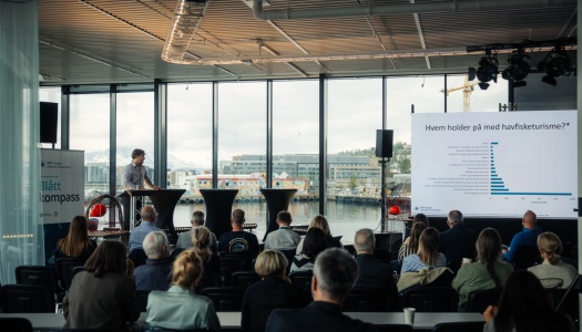 En foredragsholder presenterer for et publikum under et Blått Kompass-arrangement, med en stor skjerm som viser statistikk om havfisketurisme og utsikt mot havn og snødekte fjell gjennom vinduene bak.