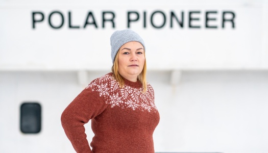En kvinne med grå strikkelue og brun ullgenser med hvitt mønster står ute foran et skip med navnet «POLAR PIONEER» i bakgrunnen.