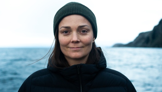 Person iført mørk jakke og grønn lue står utendørs foran et hav og fjell i bakgrunnen på en overskyet dag.