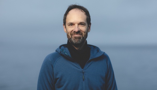 En mann står utendørs foran havet og smiler mot kameraet. Han har på seg en blå hettegenser og har kort hår og skjegg.