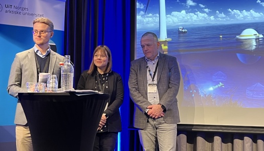 Tre personer står på en scene under et arrangement ved UiT Norges arktiske universitet; en presentasjons­skjerm bak dem viser et havmiljø med offshore vindmølle og havinstallasjoner.