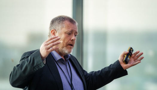 Mann med skjegg og mikrofon gestikulerer engasjert mens han holder en presentasjonsklikker.