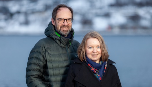 En mann og en kvinne står ute foran et vann med snødekte fjell i bakgrunnen. Mannen har briller og grønn dunjakke, kvinnen har mørk kåpe og blått skjerf med mønster.