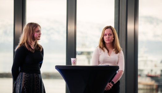 To unge kvinner står på en scene bak et høyt rundt bord under en paneldiskusjon. Begge har mikrofoner og ser konsentrerte ut. I bakgrunnen sees store vinduer med utsikt over snødekte fjell og fjord.