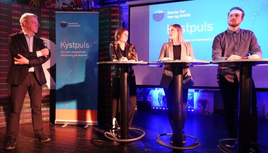 Fire personer deltar i en paneldebatt på en scene under arrangementet «Kystpuls», arrangert av Senter for hav og Arktis. En mann står med mikrofon til venstre, mens tre personer står bak høye bord foran en skjerm med arrangementets logo.