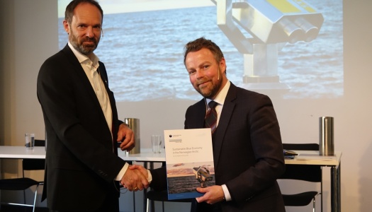 To menn i dress håndhilser mens den ene holder en rapport med tittelen «Sustainable Blue Economy in the Norwegian Arctic». Et bilde av havet vises på skjermen bak dem.