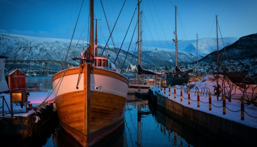 Trebåt fortøyd ved kai i vinterlys i Tromsø havn, med Tromsøbrua og snødekte fjell i bakgrunnen.