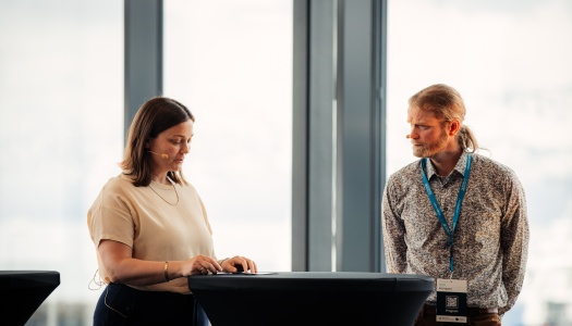 To personer deltar i en panelsamtale foran store vinduer med utsikt over et snødekt fjellandskap. Kvinnen til venstre står bak et rundt bord og ser ned på notatene sine, mens mannen til høyre ser på henne og lytter oppmerksomt. Begge har mikrofoner festet ved kinnet.