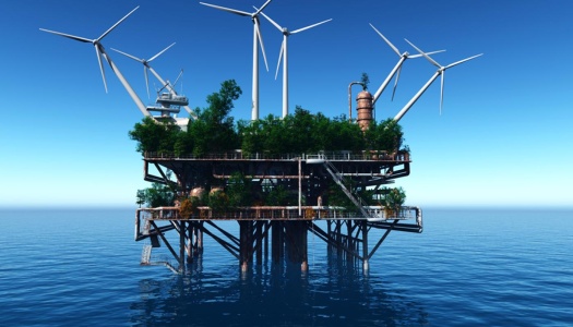 En ombygd, rusten havplattform ute på åpent hav, dekket av trær og vegetasjon, med flere store vindturbiner montert rundt strukturen under en klar blå himmel.